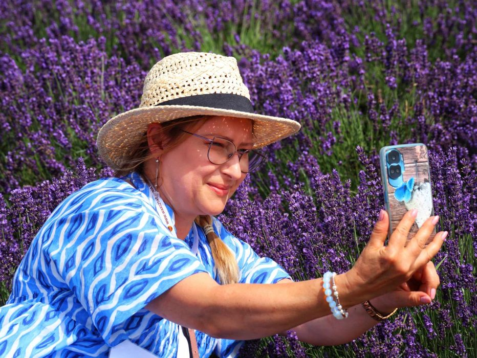 Kobieta w słomkowym kapeluszu i niebiesko-białej bluzce robi zdjęcie telefonem na tle gęstych, fioletowych krzewów lawendy podczas wycieczki