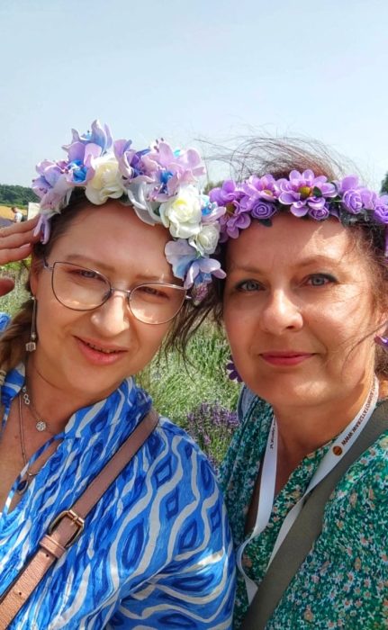 Bliskie ujęcie (selfie) dwóch uśmiechniętych kobiet w ozdobnych wiankach z kwiatów na tle pola lawendy. Monika Wojdyło (po prawej) ma na sobie fioletowy wianek i zieloną sukienkę w kwiaty