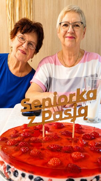 Dwie seniorki siedzą przy stole i uśmiechają się do obiektywu podczas spotkania w Warsztatowni. Na pierwszym planie znajduje się efektowne, czerwone ciasto z truskawkami i dekoracyjnym napisem „seniorka z Pasją”, a obok stoją wysoki szklany napój oraz telefon. W tle widać drewnianą ścianę, zasłony i kilka roślin doniczkowych
