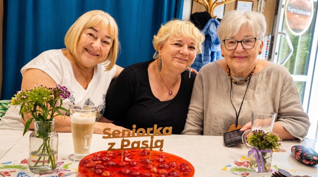 Trzy seniorki siedzą przy stole i uśmiechają się do obiektywu podczas wspólnego spotkania. Na stole widać czerwony deser lub tort z napisem „seniorka z Pasją”, małe bukiety kwiatów w szklanych wazonikach oraz napój kawowy z pianką. W tle znajduje się granatowa zasłona, wieszak na ubrania i jasne wnętrze sali