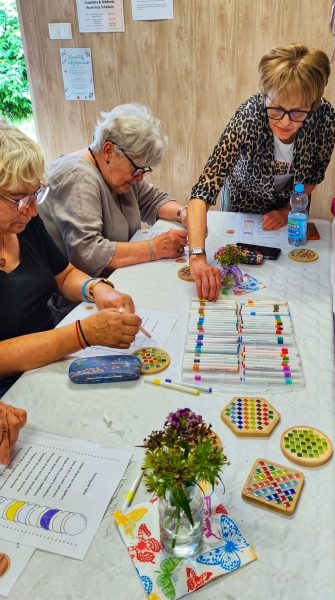 Trzy seniorki pracują przy stole podczas warsztatów w jasnym, drewnianym wnętrzu. Jedna z kobiet pokazuje wzór na kartce i wskazuje miejsce pracy, a pozostałe skupiają się na własnych zadaniach. Na stole leżą kolorowe markery, ozdobne podkładki, mały bukiet kwiatów oraz materiały warsztatowe, co nadaje scenie twórczy i uporządkowany charakter
