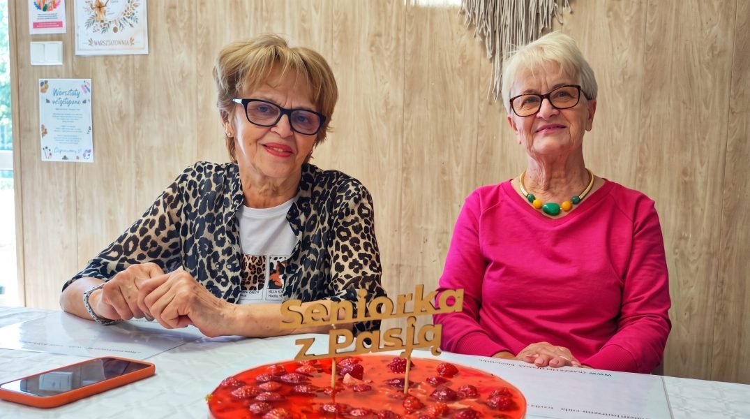 Dwie seniorki siedzą przy stole i uśmiechają się do obiektywu podczas spotkania w Warsztatowni. Przed nimi stoi dekoracyjny czerwony deser z napisem „seniorka z Pasją”, a obok znajdują się małe kwiatowe bukieciki w szklanych wazonikach oraz telefon. W tle widać jasną ścianę z drewnianą okładziną i plakaty warsztatowe