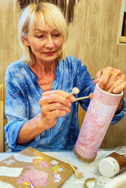 Dojrzała kobieta o blond włosach, ubrana w błękitną koszulę, w skupieniu dekoruje wysoki, różowy wazon przy użyciu pędzla gąbkowego