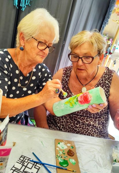 Dwie dojrzałe kobiety w okularach w skupieniu malują wysoki, błękitny wazon ozdobiony motywem różowej róży