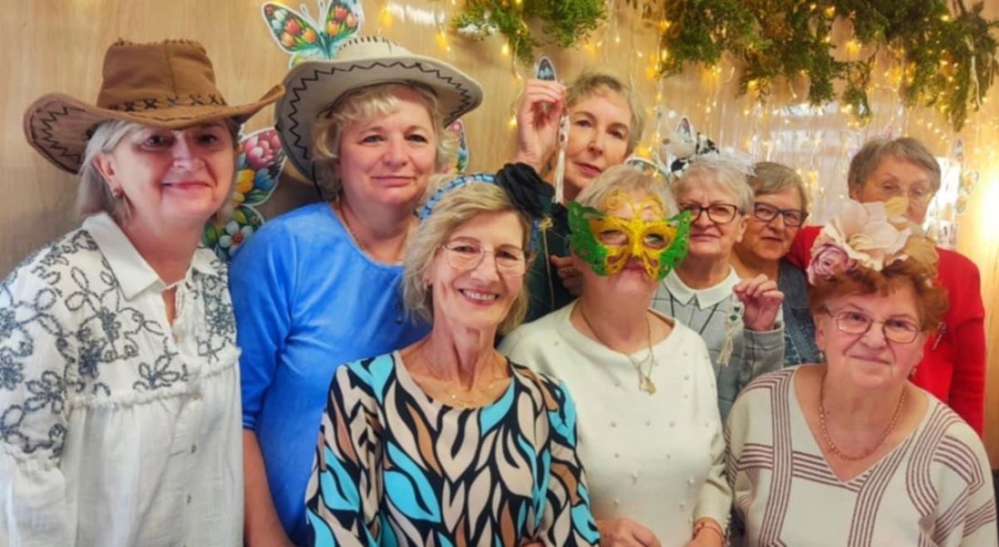 Grupa uśmiechniętych starszych kobiet pozuje razem do zdjęcia; niektóre z nich mają na sobie kolorowe kapelusze, opaski i maskę karnawałową