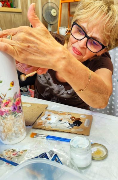 Dojrzała kobieta w okularach i brązowej koszulce w skupieniu maluje biały wazon z motywem kwiatowym, trzymając pędzel gąbkowy