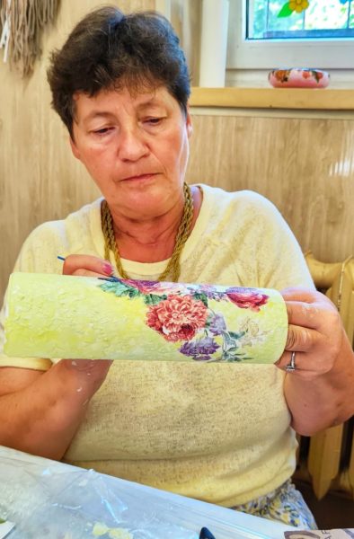 Dojrzała kobieta w żółtej bluzce w skupieniu trzyma poziomo wysoki wazon z motywem różowych i fioletowych kwiatów, pracując nad jego wykończeniem