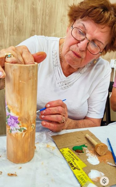 Dojrzała kobieta w okularach i białej bluzce w skupieniu maluje wysoki, drewniany lub ceramiczny wazon w kolorze miodowym, ozdobiony fioletowym kwiatem
