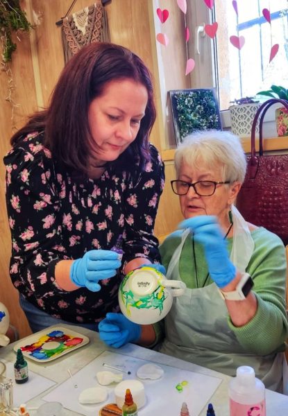 Instruktorka Monika Wojdyło w kwiecistej bluzce i niebieskich rękawiczkach pomaga dojrzałej kobiecie ozdabiać białą filiżankę kolorowymi farbami