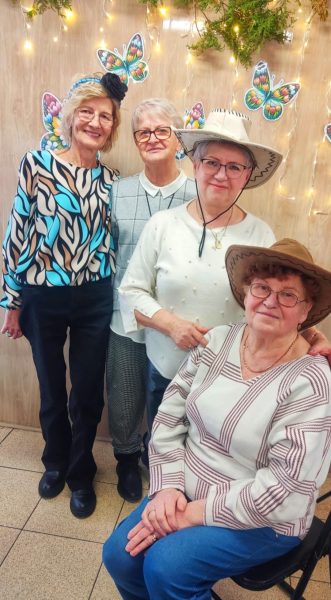 Cztery uśmiechnięte kobiety w starszym wieku pozują razem do zdjęcia; dwie z nich mają na sobie kapelusze kowbojskie, a jedna ozdobną opaskę z kwiatem
