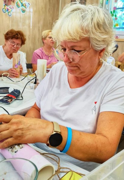 Dojrzała kobieta w okularach i białej koszulce z małym czerwonym sercem na piersi, w skupieniu pracuje przy stole warsztatowym