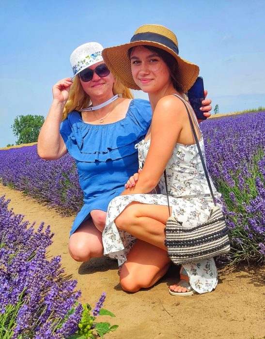 Dwie uśmiechnięte kobiety pozują, kucając pośród rzędów fioletowej lawendy. Jedna ma na sobie niebieską sukienkę i biały kapelusz z kwiatowym wzorem, a druga białą sukienkę w kwiaty i duży słomkowy kapelusz