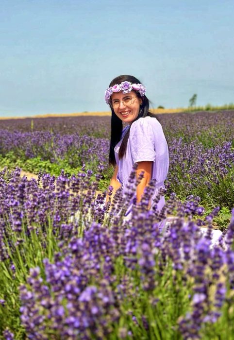 Uśmiechnięta kobieta w liliowej sukience i wianku z jasnofioletowych kwiatów pozuje, siedząc pośród kwitnącej lawendy pod błękitnym niebem