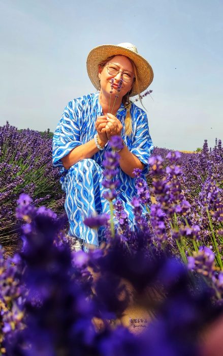 Kobieta w niebiesko-białej wzorzystej sukience i słomkowym kapeluszu kuca w gęstym rzędzie kwitnącej lawendy, trzymając kilka gałązek kwiatów przy twarzy i uśmiechając się