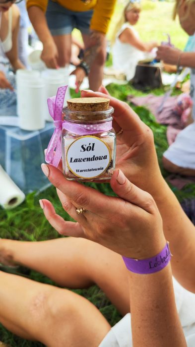 Zbliżenie na dłonie trzymające szklany słoiczek wypełniony różową solą z dodatkiem suszonej lawendy. Słoik ma korkowe zamknięcie, różową kokardkę i etykietę „Sól Lawendowa Warsztatownia”