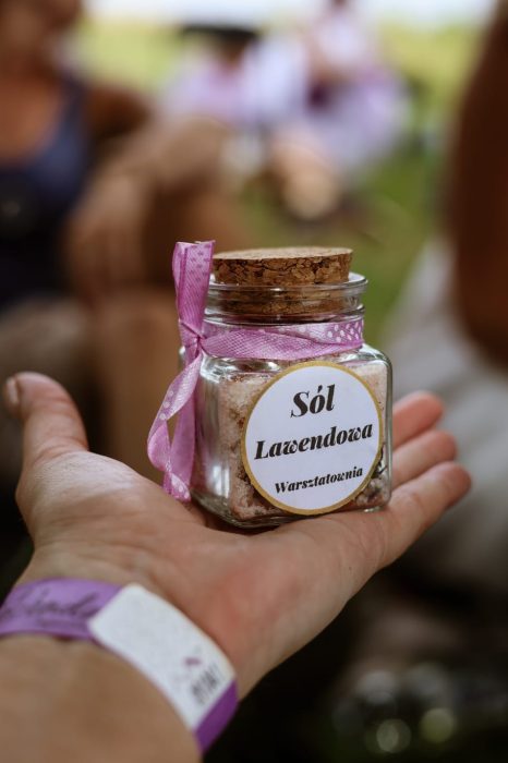 Zbliżenie na dłoń trzymającą mały szklany słoiczek z solą lawendową, ozdobiony różową wstążką i etykietą z napisem „Sól Lawendowa Warsztatownia”