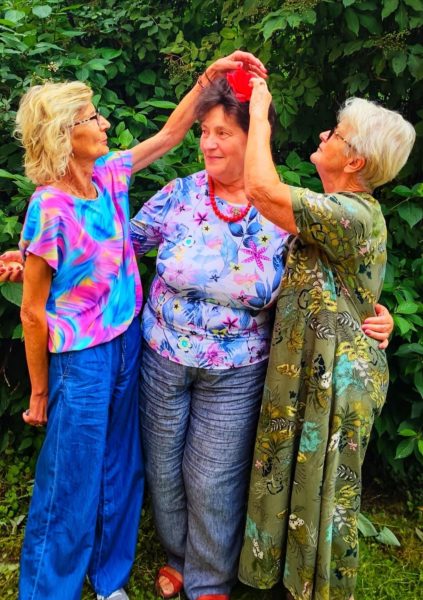 Trzy uśmiechnięte kobiety w kolorowych ubraniach stoją na tle zieleni; dwie z nich pomagają trzeciej założyć czerwoną ozdobę na włosy