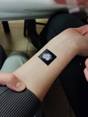 Zbliżenie na przedramię dziecka z małym, tymczasowym tatuażem lub naklejką w formie czarnego kwadratu z delikatną grafiką pośrodku. Dłoń osoby dorosłej lekko podtrzymuje rękę dziecka. W tle widać wnętrze z krzesłami i stołem, co sugeruje zabawę lub uroczystość urodzinową