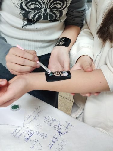 Zbliżenie na przedramię dziecka z małym, tymczasowym tatuażem lub naklejką w formie czarnego kwadratu z delikatną grafiką pośrodku. Dłoń osoby dorosłej lekko podtrzymuje rękę dziecka. W tle widać wnętrze z krzesłami i stołem, co sugeruje zabawę lub uroczystość urodzinową