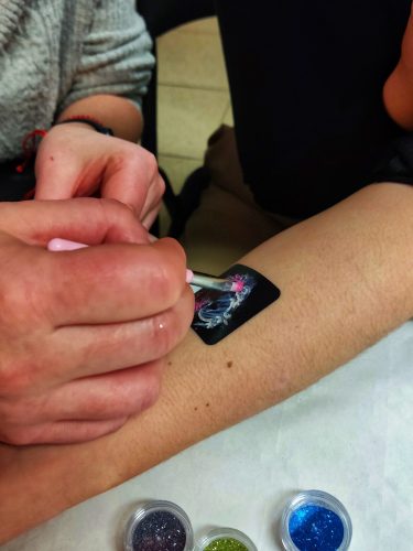 Zbliżenie na przedramię dziecka z małym, tymczasowym tatuażem lub naklejką w formie czarnego kwadratu z delikatną grafiką pośrodku. Dłoń osoby dorosłej lekko podtrzymuje rękę dziecka. W tle widać wnętrze z krzesłami i stołem, co sugeruje zabawę lub uroczystość urodzinową
