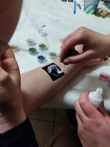 Zbliżenie na przedramię dziecka z małym, tymczasowym tatuażem lub naklejką w formie czarnego kwadratu z delikatną grafiką pośrodku. Dłoń osoby dorosłej lekko podtrzymuje rękę dziecka. W tle widać wnętrze z krzesłami i stołem, co sugeruje zabawę lub uroczystość urodzinową