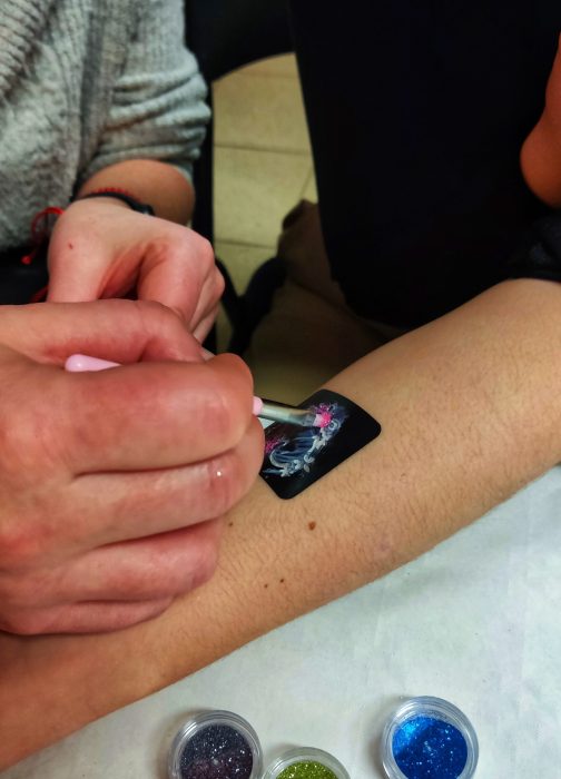 Zbliżenie na dłoń animatora nakładającą różowy brokat pędzelkiem na czarny szablon tatuażu na przedramieniu dziecka. Na dole widoczne słoiczki z kolorowym brokatem: fioletowym, zielonym i niebieskim