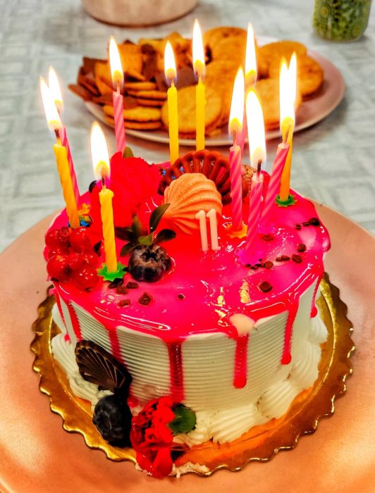 Kolorowy tort urodzinowy z różową polewą, udekorowany świeżymi owocami (porzeczki, borówki), kwiatami i bezami. Na torcie płonie 11 żółtych i różowych świeczek, a obok widnieje świeczka w kształcie liczby 11