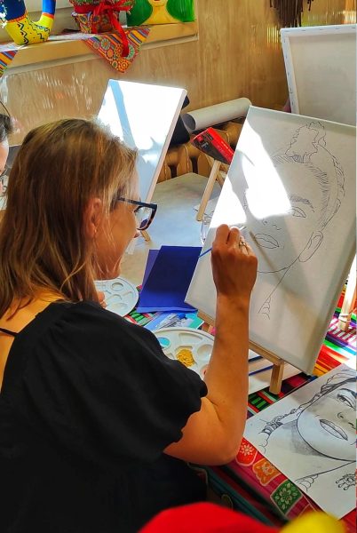 Uczestniczka warsztatów z długimi blond włosami i okularami, skupiona na malowaniu konturu twarzy Fridy Kahlo na płótnie, z paletą farb i dekoracjami meksykańskimi w tle