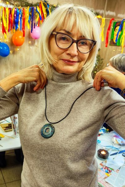 Portret uśmiechniętej kobiety w beżowym golfie i okularach, która obiema rękami trzyma rzemyk zawieszonego na szyi wisiora. Wisior jest okrągły, wykonany techniką wikliny papierowej w odcieniach zieleni, błękitu i czerni. W tle widać kolorowe balony, serpentyny oraz stół warsztatowy z materiałami plastycznymi