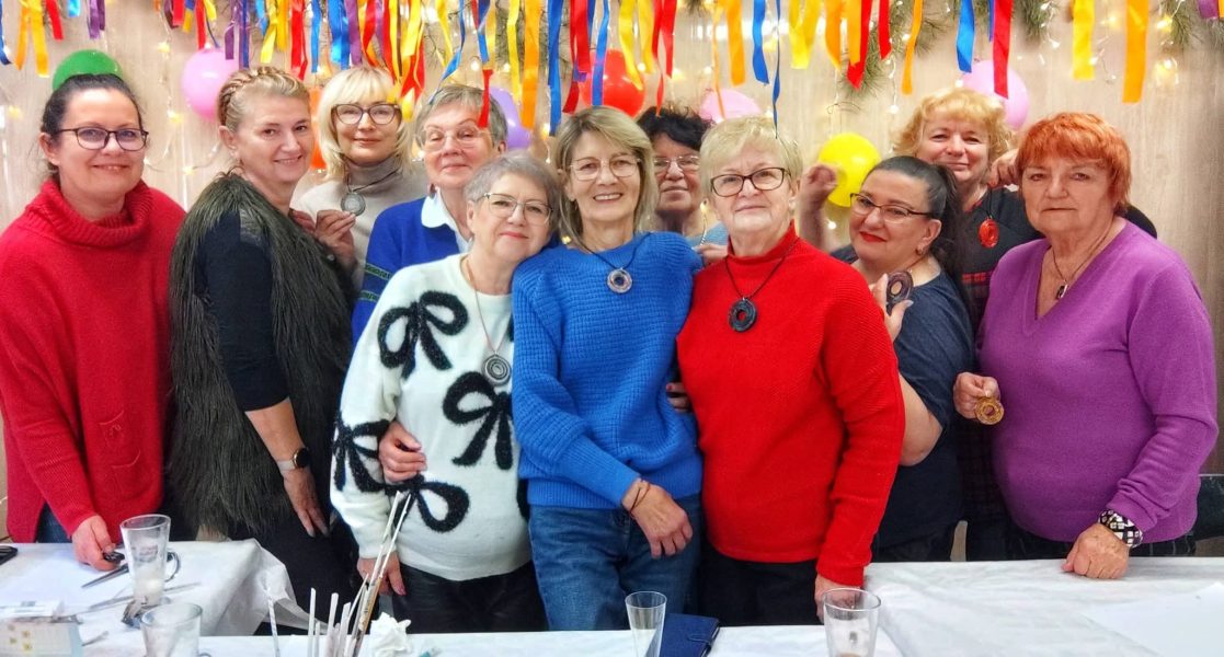 Grupa dziesięciu uśmiechniętych kobiet pozuje do wspólnego zdjęcia po warsztatach rękodzielniczych. Uczestniczki dumnie prezentują na szyjach i w dłoniach własnoręcznie wykonane, okrągłe wisiory z wikliny papierowej w kolorach złotym, granatowym i czerwonym. Tło zdobią kolorowe balony, jasne lampki i wielobarwne serpentyny, tworzące radosną, odświętną atmosferę
