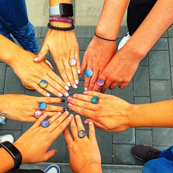 Siedem par kobiecych dłoni ułożonych w okręgu, prezentujących kolorowe, lśniące pierścionki wykonane z żywicy UV. Każdy pierścionek ma unikatową kompozycję barw – od morskich błękitów i brokatowych fioletów po opalizujące biele. Tło stanowi szara kostka brukowa