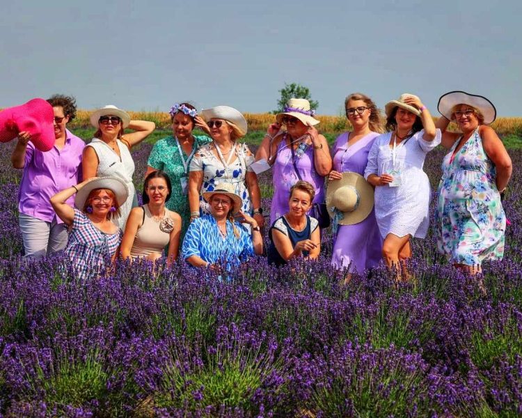 Grupa kilkunastu uśmiechniętych kobiet w letnich stylizacjach i kapeluszach pozuje wspólnie pośród rzędów kwitnącej lawendy pod błękitnym niebem