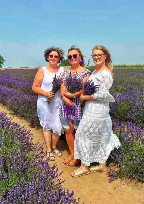 Trzy uśmiechnięte kobiety w letnich stylizacjach stoją na ścieżce pośród rzędów fioletowej lawendy. Każda z nich trzyma w dłoniach bukiet świeżo zerwanej lawendy