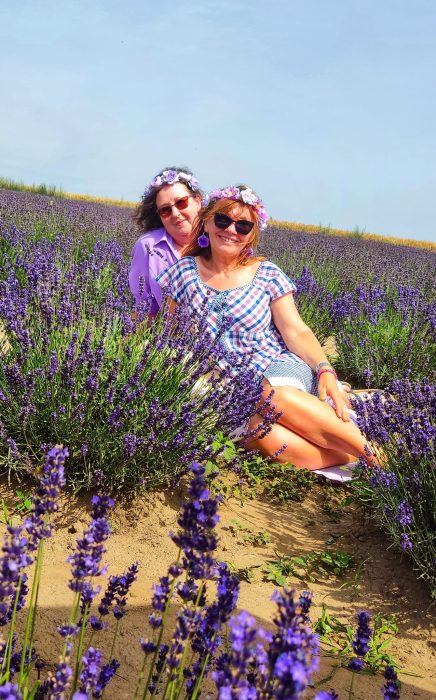 Dwie uśmiechnięte kobiety siedzą pośród rzędów fioletowej lawendy pod błękitnym niebem. Obie mają na sobie ozdobne wianki z fioletowych kwiatów i letnie stylizacje