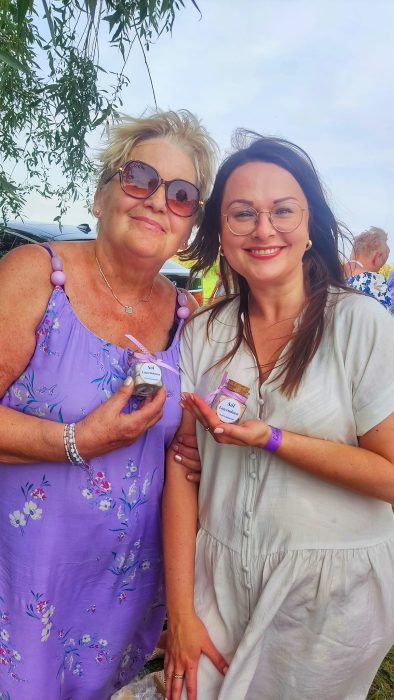 Dwie uśmiechnięte kobiety w letnich stylizacjach (fioletowa sukienka w kwiaty oraz biała sukienka) trzymają na dłoniach gotowe słoiczki soli lawendowej z różowymi kokardkami