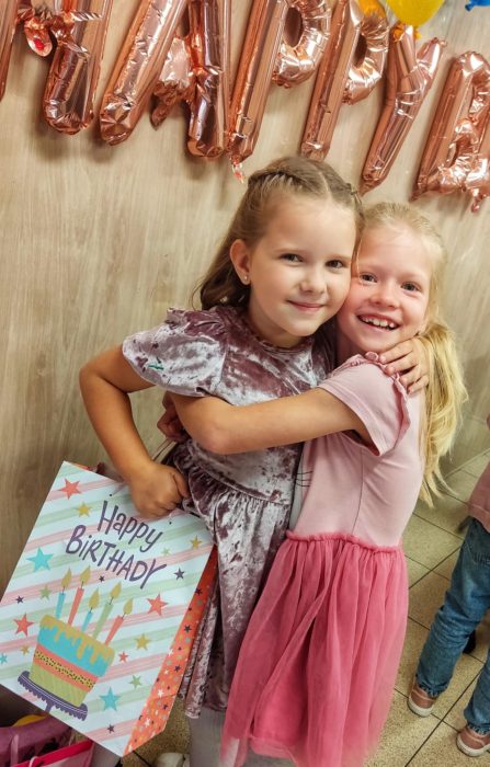 Dwie uśmiechnięte dziewczynki tulące się do siebie podczas przyjęcia urodzinowego. Jedna z nich trzyma kolorową torebkę prezentową z napisem „Happy Birthday”, w tle widoczne różowozłote balony tworzące napis „HAPPY”