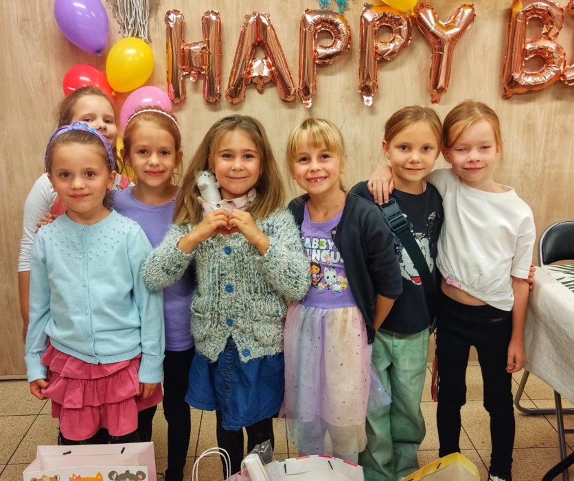 Grupa siedmiu uśmiechniętych dziewczynek pozuje na tle drewnianej ściany z napisem „HAPPY BIRTHDAY” z różowozłotych balonów oraz kolorowymi balonami: fioletowym, żółtym i czerwonym