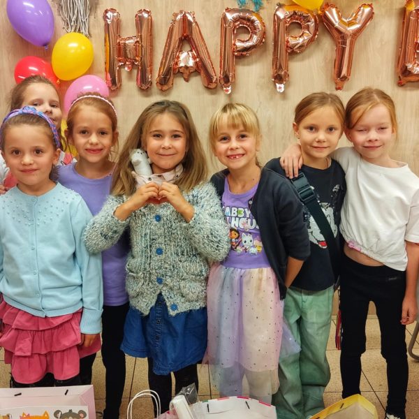 Grupa siedmiu uśmiechniętych dziewczynek pozuje na tle drewnianej ściany z napisem „HAPPY BIRTHDAY” z różowozłotych balonów oraz kolorowymi balonami: fioletowym, żółtym i czerwonym