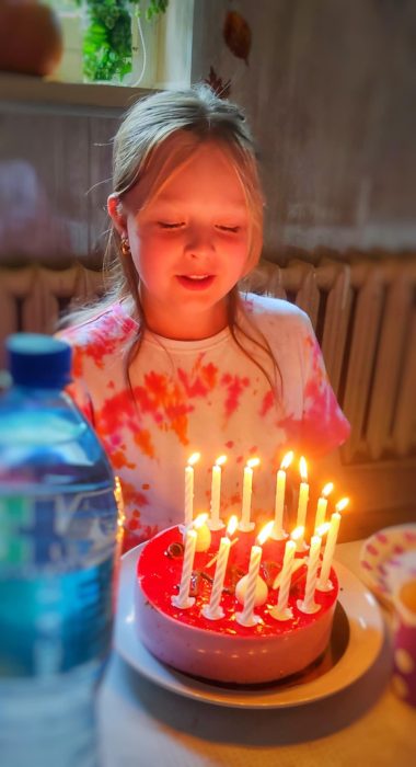 Dziewczynka w kolorowej koszulce tye-dye z uśmiechem i zamkniętymi oczami pochyla się nad różowym tortem urodzinowym z wieloma płonącymi świeczkami. Ciepłe światło świec oświetla twarz dziecka