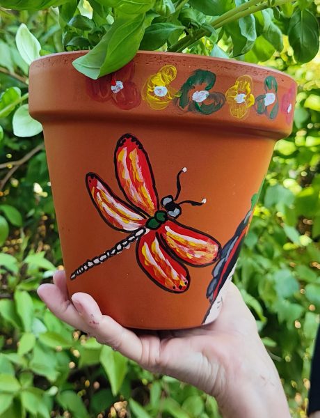 Pomarańczowa gliniana doniczka ręcznie pomalowana ważką – skrzydła w odcieniach czerwieni i żółci, kwiatowe akcenty i zielone liście, trzymana w dłoni na tle roślinności