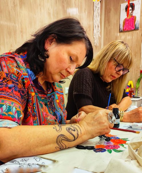 Dwie kobiety skupione na malowaniu białych bawełnianych toreb farbami akrylowymi podczas warsztatów w Warsztatowni, z widocznymi kwiatowymi motywami, pędzlami i dekoracjami w tle