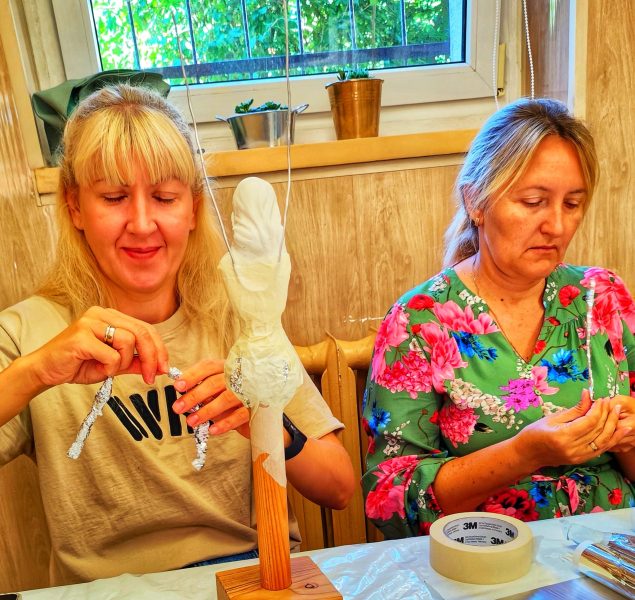 Dwie kobiety w skupieniu pracują nad stelażem rzeźby z użyciem folii i taśmy podczas warsztatów technicznych w Warsztatowni