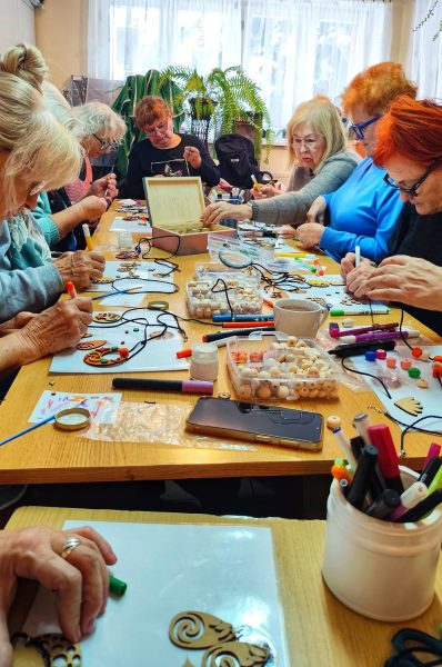 Duża grupa seniorek w skupieniu tworzy biżuterię z koralików przy wspólnym stole podczas warsztatów Warsztatowni