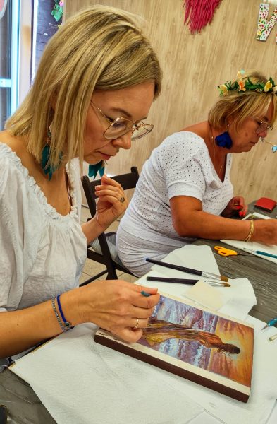 Kobieta w okularach w skupieniu maluje pędzlem obraz na drewnie podczas warsztatów artystycznych w Warsztatowni