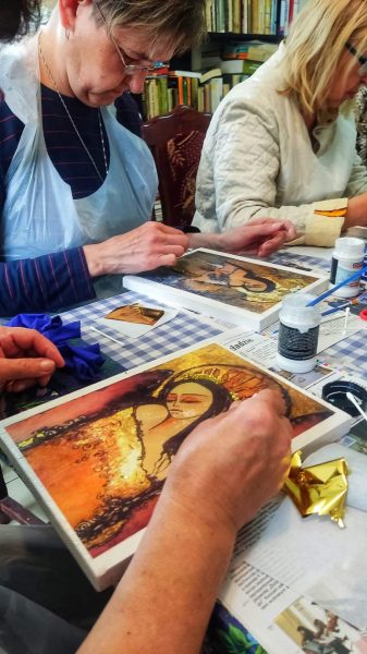 Zbliżenie na ręce uczestniczki warsztatów nakładającej płatki złota na obraz w Warsztatowni; w tle inna osoba pracuje nad podobnym projektem