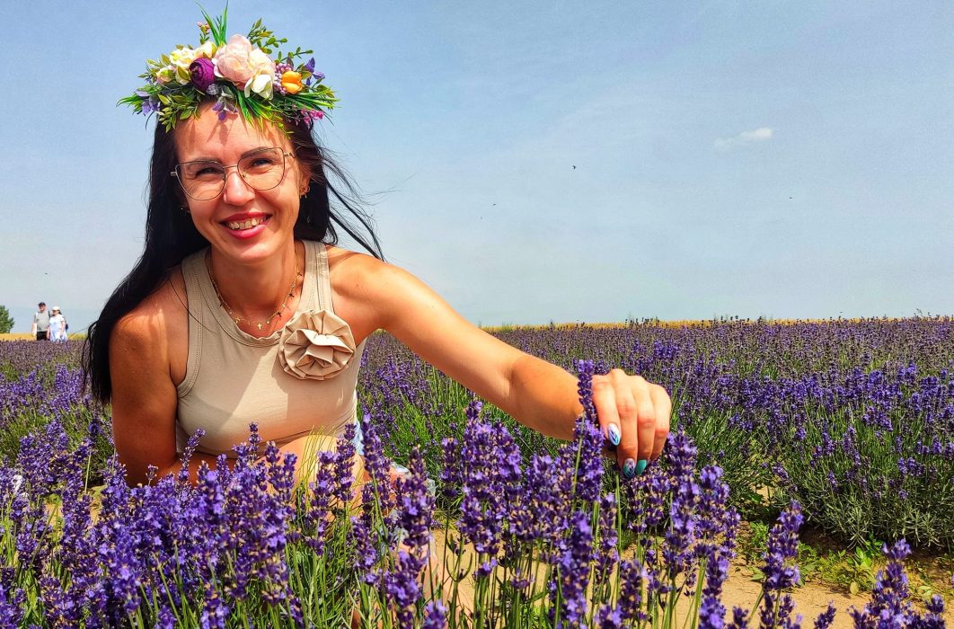 Bliskie ujęcie uśmiechniętej kobiety w okularach i kolorowym wianku z kwiatów, która kuca na polu i delikatnie dotyka dłonią krzewu kwitnącej, fioletowej lawendy