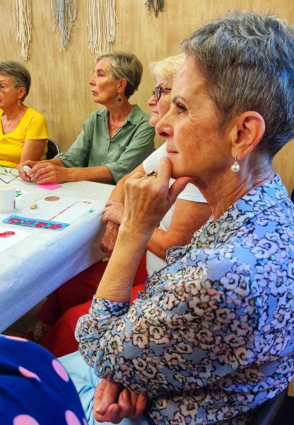 Profil dojrzałej kobiety z krótkimi siwymi włosami i perłowym kolczykiem, która w zamyśleniu trzyma dłoń przy brodzie. W tle widać inne uczestniczki warsztatów siedzące przy wspólnym stole