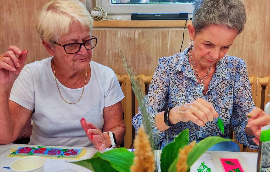 Dwie dojrzałe kobiety siedzą obok siebie przy warsztatowym stole. Jedna z nich, w okularach i białym t-shircie, z uśmiechem obserwuje pracę koleżanki w błękitnej bluzce, która precyzyjnie maluje zielony listek