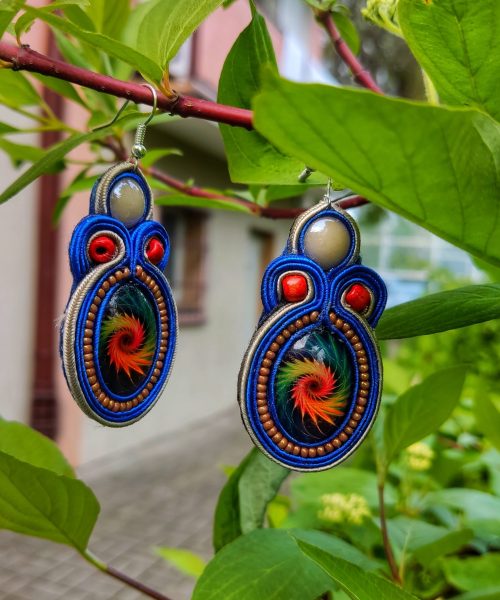 Para kolczyków sutasz w kolorze intensywnego błękitu, zawieszona na zielonej gałązce. Kolczyki zdobią jasne kaboszony u góry oraz owalne oczka z motywem ognistopomarańczowej spirali, otoczone drobnymi miedzianymi koralikami