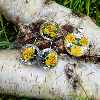 Zestaw biżuterii z żywicy UV: para okrągłych kolczyków, wisior i pierścionek, ułożone na pniu brzozy. W przezroczystej żywicy zatopiono żółte bratki z ciemnymi środkami oraz dużą ilość srebrnego, mieniącego się brokatu